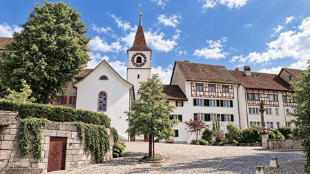Historischer Dorfplatz von Regensberg mit weisser Kirche und markantem Uhrturm, traditionellen Fachwerkhäusern mit roten Ziegeldächern und grünen Fensterläden. Der gepflasterte Platz und die alten Natursteinmauern unterstreichen den mittelalterlichen Charme des Zürcher Landstädtchens.