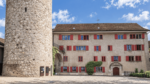Historischer Wehrturm von Regensberg mit angrenzendem Gebäude und roten Fensterläden. Der mittelalterliche Innenhof ist mit hellem Kies gepflastert und zeigt die historische Architektur des Zürcher Landstädtchens.