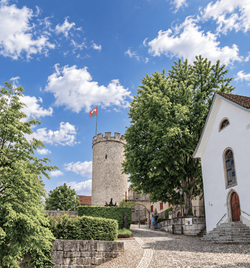 Historischer Wehrturm von Regensberg mit wehender Fahne, umgeben von grünen Bäumen, einer weissen Kapelle und einem gepflasterten Weg im mittelalterlichen Ortskern.