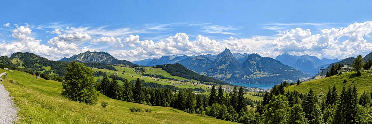 Panoramablick von Amden am Mattstockfuss über grüne Alpenwiesen und verstreute Häuser auf die umliegende Bergwelt und den Walensee bei sommerlichem Wetter.