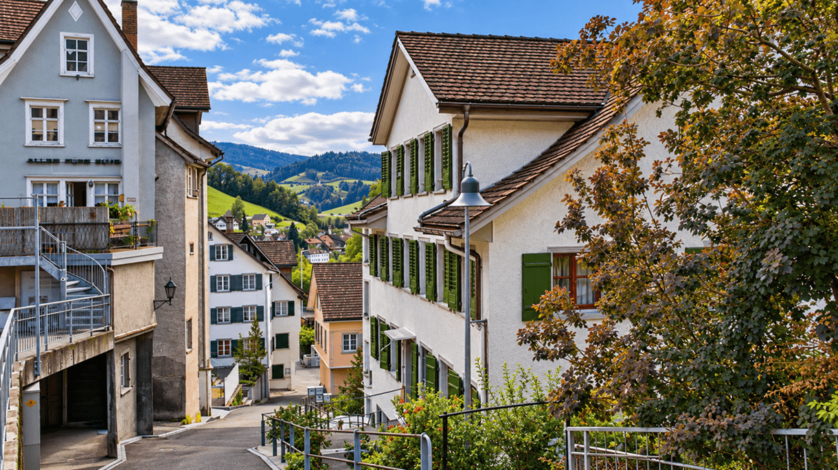 Schweizer Dorfgasse mit traditionellen Häusern, grünen Fensterläden und herbstlich gefärbten Bäumen. Im Hintergrund erstrecken sich grüne Hügel und bewaldete Höhen unter blauem Himmel.
