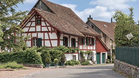 Historisches Fachwerkhaus «Chräzli» in Regensberg mit weiss verputzten Wänden, roten Holzbalken, grünen Fensterläden und traditionellem Ziegeldach. Das gepflegte Gebäude mit üppiger Begrünung liegt an einer ruhigen Strasse im mittelalterlichen Ortskern.